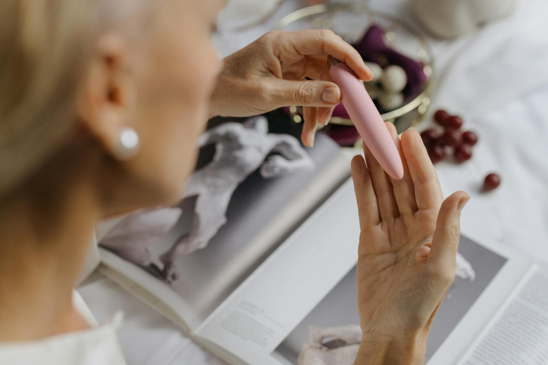 Close-up of a woman holding a pink vibrator with art books in the background, representing intimate exploration with a partner.